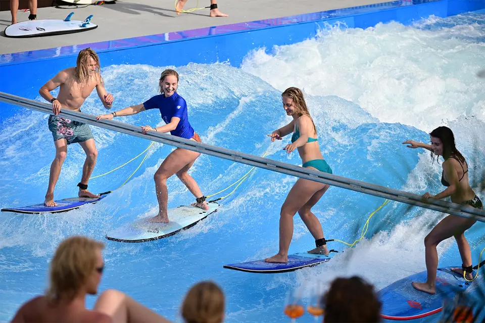 Vier Personen surfen auf der stehenden Wellen in Freiburg und balancieren auf Brettern. Andere schauen von der Seite zu.