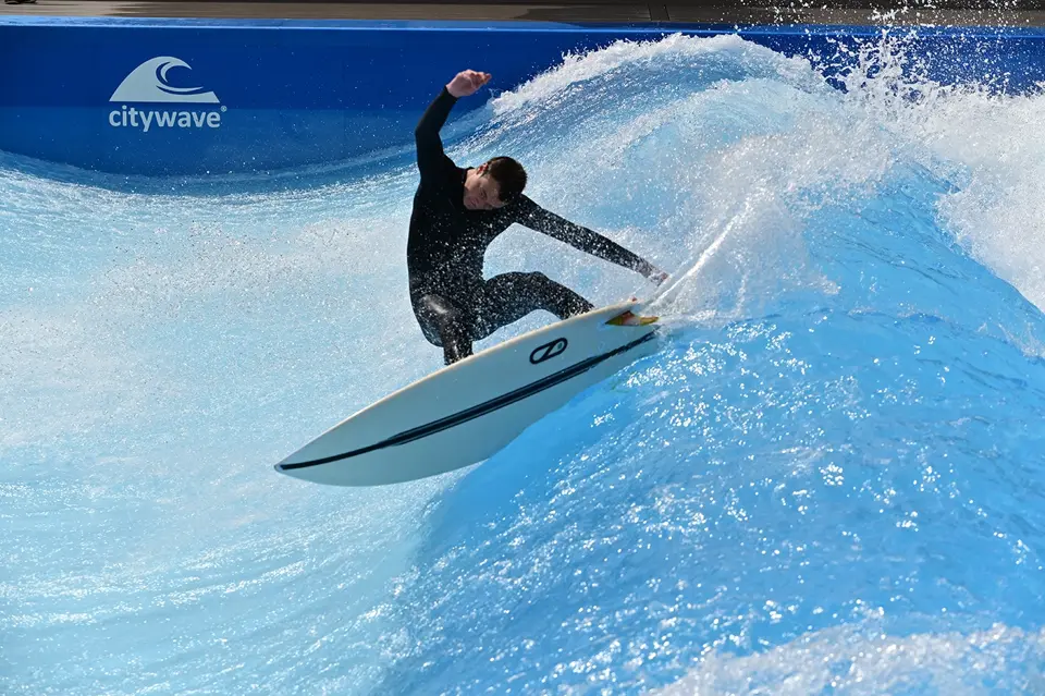 Ein Surfer in einem schwarzen Neoprenanzug reitet auf der Welle von irie surf in einem künstlichen Surfbecken und führt ein Manöver durch, bei dem das Wasser herumspritzt.
